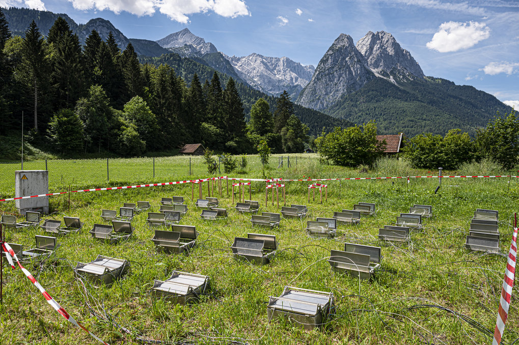 
Zu sehen ist eine Grasfläche vor dem Hintergrund der Zugspitze und des Waxensteine. Auf der Grasfäche ist eine großflächhige wissenschaftliche Versuchsanordnung zu sehen die aus dynamischen Messkammern für die Klimagasmessungen besteht.